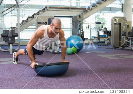 Man performing balancing push-up on Bosu ball on purple gym carpet with weight machines, copy space Man performing balancing push-up on Bosu ball on purple gym carpet with weight machines, copy space 135408224