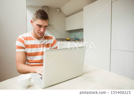 Man sitting at kitchen table focusing on silver laptop with mug, fruit bowl and water bottle Man sitting at kitchen table focusing on silver laptop with mug, fruit bowl and water bottle 135408225