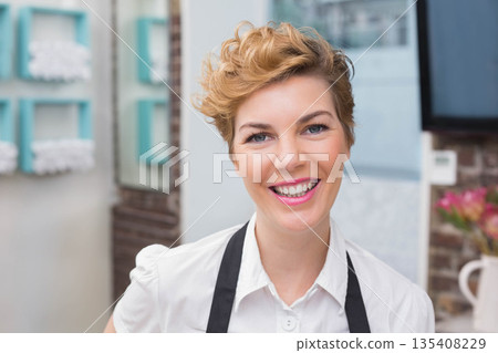 Woman barista smiling while standing behind teal shelves and menu board wearing black apron Woman barista smiling while standing behind teal shelves and menu board wearing black apron 135408229