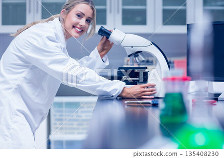 Female scientist leaning over lab bench adjusting microscope stage handling reagent bottles 135408230