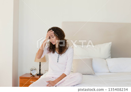 Woman wearing pajamas sitting on bed holding forehead with nightstand lamp clock glass 135408234