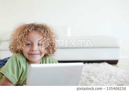 Boy lying on white shag rug in living room using silver laptop beside white low-backed sofa 135408238