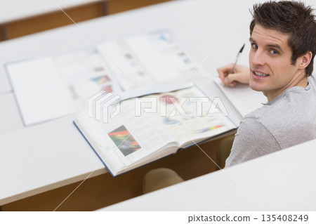 Male student writing notes with pen looking toward camera at lecture desk with textbook, copy space 135408249
