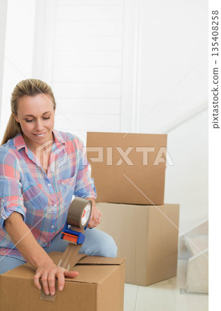 Woman kneeling on floor near staircase at home taping cardboard box with tape dispenser 135408258