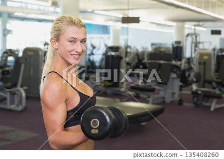 Woman lifting dumbbell at shoulder level in gym wearing sportswear with weight machines and mirrors Woman lifting dumbbell at shoulder level in gym wearing sportswear with weight machines and mirrors 135408260