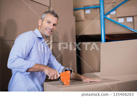 Middle-aged man using orange tape dispenser sealing cardboard boxes at warehouse with blue shelving Middle-aged man using orange tape dispenser sealing cardboard boxes at warehouse with blue shelving 135408263