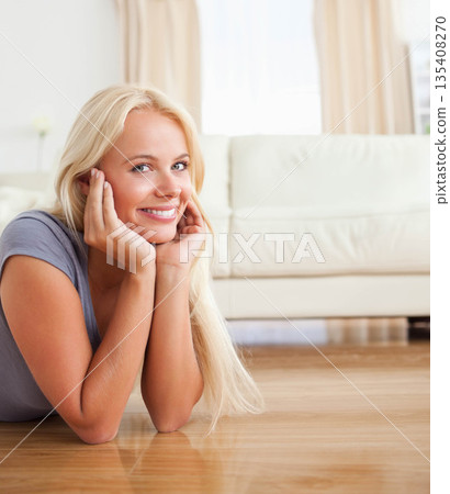 Woman lying on polished wooden floor in bright living room smiling at camera near white couch 135408270