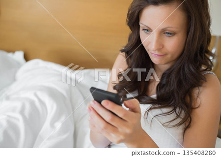 Woman sitting on bed holding smartphone and reading messages in bedroom, copy space 135408280