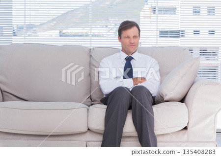 Mature man sitting in office lounge on sofa crossing arms while gazing through blinds Mature man sitting in office lounge on sofa crossing arms while gazing through blinds 135408283