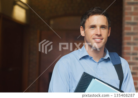 Man in his thirties smiling while holding black binder folder and shoulder bag in building corridor 135408287