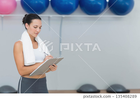 Female trainer surveying balls, balance trainers in studio holding clipboard with towel, copy space 135408289