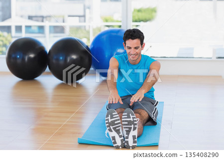 Man in late twenties stretching forward on blue exercise mat in bright studio with stability balls 135408290