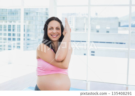 Pregnant woman standing on blue yoga mat and holding yoga pose in studio overlooking city skyline Pregnant woman standing on blue yoga mat and holding yoga pose in studio overlooking city skyline 135408291