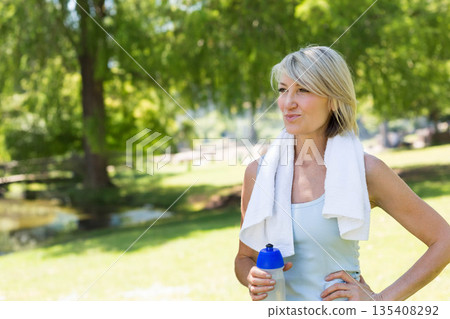 Senior woman standing in sunlit park beside pond holding water bottle wearing tank top, copy space Senior woman standing in sunlit park beside pond holding water bottle wearing tank top, copy space 135408292