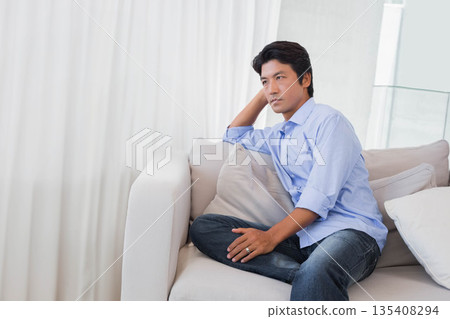 Mid adult Japanese man leaning back on light-colored sofa in bright living room with sheer curtains 135408294