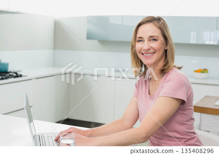 Woman smiling at camera while working on laptop at kitchen counter with fruit bowl, copy space 135408296
