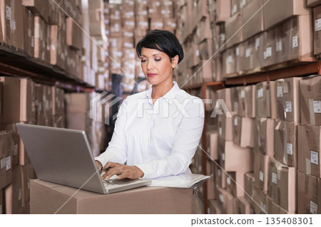 Asian woman wearing white shirt working in warehouse scanning barcodes on boxes using silver laptop Asian woman wearing white shirt working in warehouse scanning barcodes on boxes using silver laptop 135408301