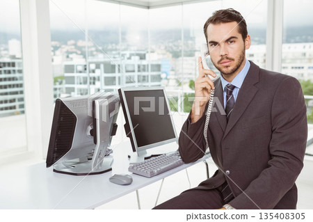 Man in suit holding telephone handset and operating dual monitors at office desk 135408305
