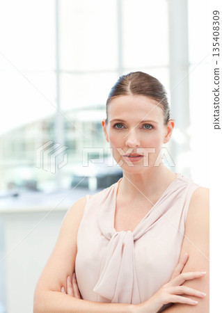 Mid adult woman standing in sleeveless blouse with crossed arms in office lobby with bright windows 135408309