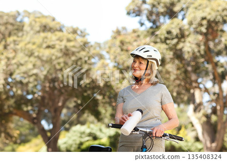 Senior woman pausing beside bicycle at park wearing white helmet holding water bottle, copy space Senior woman pausing beside bicycle at park wearing white helmet holding water bottle, copy space 135408324