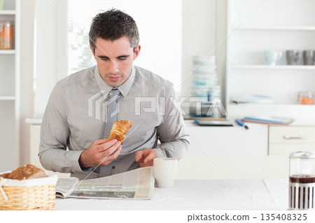 Business professional man holding croissant and reading newspaper at kitchen table with coffee mug Business professional man holding croissant and reading newspaper at kitchen table with coffee mug 135408325