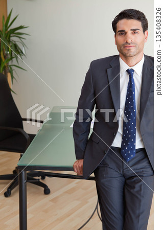 Businessman wearing suit leaning against glass-topped desk with papers at office, copy space Businessman wearing suit leaning against glass-topped desk with papers at office, copy space 135408326