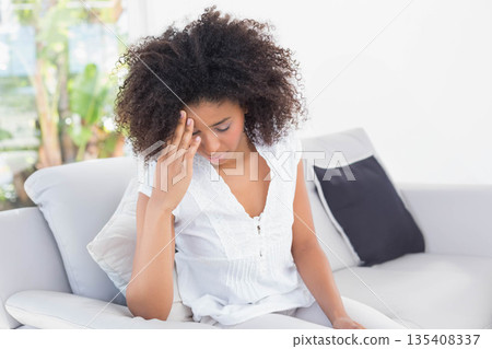 African American woman sitting on white sofa in living room holding head under window with plants African American woman sitting on white sofa in living room holding head under window with plants 135408337