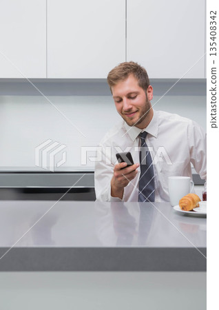 Businessman checking smartphone sitting at kitchen counter with coffee mug and croissant copy space Businessman checking smartphone sitting at kitchen counter with coffee mug and croissant copy space 135408342