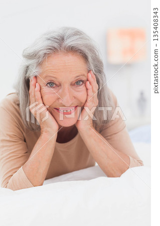 Senior woman lying on white linens in bedroom, smiling gently at bedside lamp, cupping face Senior woman lying on white linens in bedroom, smiling gently at bedside lamp, cupping face 135408343