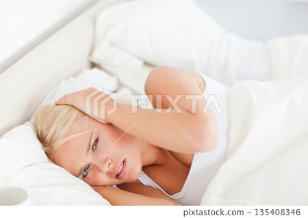 Woman lying on bed with white sheets and having headache in bright bedroom with mug beside 135408346