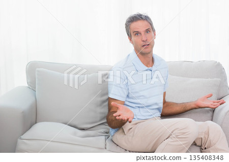 Man raising palms asking question seated on sofa in living room with cushions, curtains, copy space 135408348
