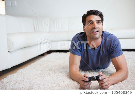 Mid adult man lying in living room on white shag rug holding black controller, copy space Mid adult man lying in living room on white shag rug holding black controller, copy space 135408349