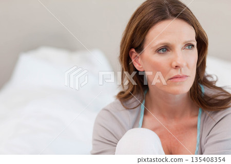 middle-aged woman sitting on bed looking toward right in bedroom with white linens, copy space 135408354