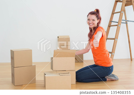 Woman kneeling in empty room near boxes and wooden ladder packing and giving thumbsup, copy space Woman kneeling in empty room near boxes and wooden ladder packing and giving thumbsup, copy space 135408355