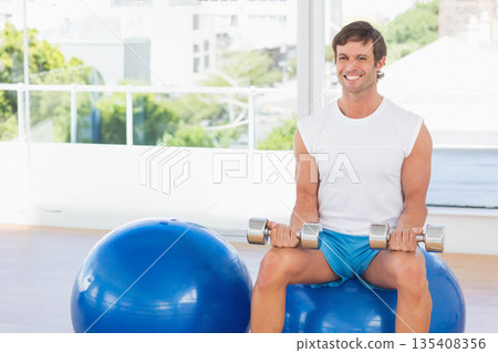 Man wearing sportswear lifting dumbbells while sitting on stability ball in studio, copy space 135408356