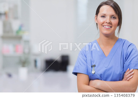 Female nurse in blue scrubs smiling while standing wearing ID badge in examination room, copy space 135408379