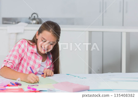 Girl aged ten sitting at home kitchen table drawing on green paper with markers, copy space 135408382
