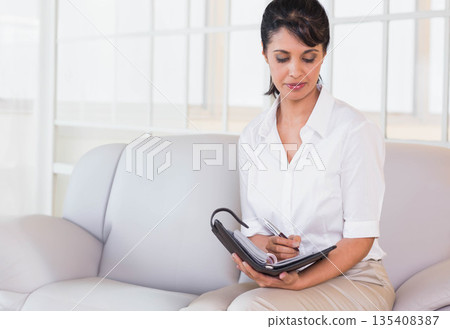 Mid adult woman writing in ring-bound planner with pen on sofa in office lounge, copy space Mid adult woman writing in ring-bound planner with pen on sofa in office lounge, copy space 135408387