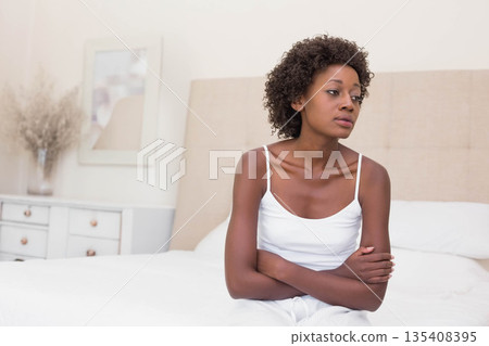 African American woman sitting on edge of unmade bed in home bedroom beside dresser with vase African American woman sitting on edge of unmade bed in home bedroom beside dresser with vase 135408395