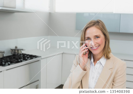Middle-aged woman wearing blazer talking on smartphone in home kitchen by stove, copy space 135408398