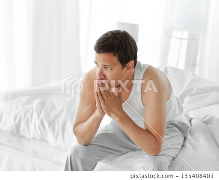 Man sitting on bed edge leaning forward with hands on mouth in bedroom with white linens 135408401