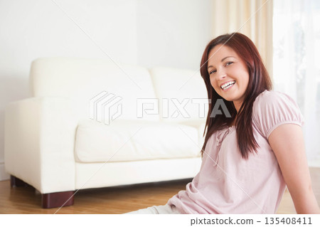 Woman sitting on wooden floor next to white sofa in living room under sheer curtains smiling 135408411