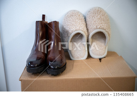 Cozy home still life with leather ankle boots and soft wool slippers on cardboard storage box Cozy home still life with leather ankle boots and soft wool slippers on cardboard storage box 135408665