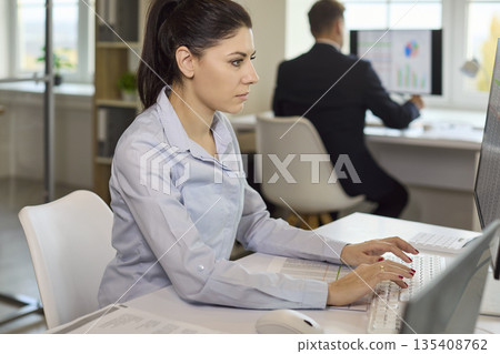 Focused office analyst typing financial data on computer while colleague works Focused office analyst typing financial data on computer while colleague works 135408762