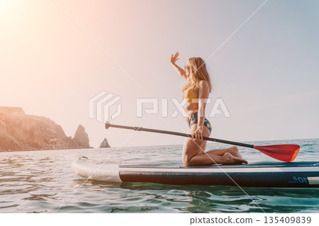 Woman Paddleboarding Ocean Coastline - A woman in a bikini kneels on a paddleboard in the ocean, holding a paddle and looking towards a cliff in the distance. Woman Paddleboarding Ocean Coastline - A woman in a bikini kneels on a paddleboard in the ocean, holding a paddle and looking towards a cliff in the distance. 135409839