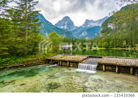 Famous green lake Schiederweiher near village Hinterstoder. Small weir and dam on lake with flowing water. Big Austrian mountains in background. Soft and magical colors in pure nature. 135410366