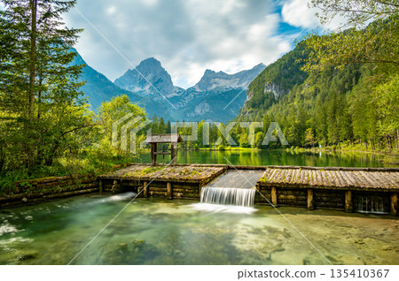Famous green lake Schiederweiher near village Hinterstoder. Small weir and dam on lake with flowing water. Big Austrian mountains in background. Soft and magical colors in pure nature. Famous green lake Schiederweiher near village Hinterstoder. Small weir and dam on lake with flowing water. Big Austrian mountains in background. Soft and magical colors in pure nature. 135410367
