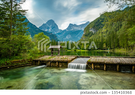 Famous green lake Schiederweiher near village Hinterstoder. Small weir and dam on lake with flowing water. Big Austrian mountains in background. Soft and magical colors in pure nature. Famous green lake Schiederweiher near village Hinterstoder. Small weir and dam on lake with flowing water. Big Austrian mountains in background. Soft and magical colors in pure nature. 135410368