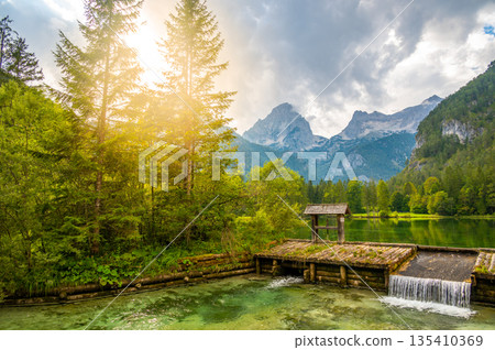 Famous green lake Schiederweiher near village Hinterstoder. Small weir and dam on lake with flowing water. Big Austrian mountains in background. Soft and magical colors in pure nature. 135410369