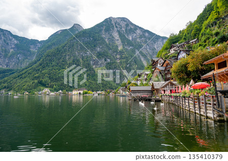 Hallstatt Village and Hallstatter See lake in Austria. Scenery with famous old church near the lake. Clouds and mist over the mountains in background. Famous tourist destination. Hallstatt Village and Hallstatter See lake in Austria. Scenery with famous old church near the lake. Clouds and mist over the mountains in background. Famous tourist destination. 135410379